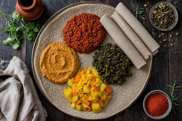Äthiopische Fastentafel von oben: eine große Injera-Fladenbrotplatte, belegt mit Häufchen aus rotem Misir-Wot-Linseneintopf, cremigem Shiro, dunkelgrünem Gomen-Grünkohl und gelbem Atakilt-Wat-Gemüse, dazu gerollte Injera-Streifen am Rand