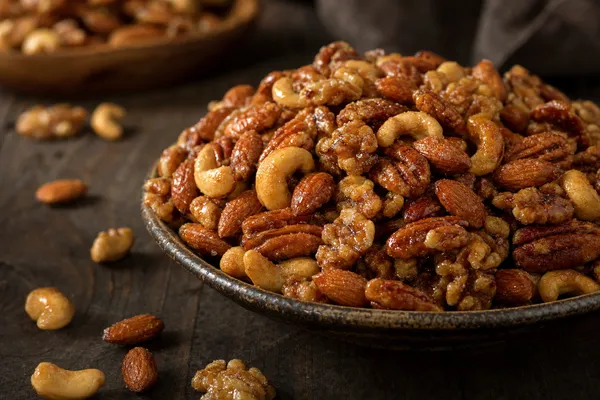Eine Schale glänzend ahornglasierter gemischter Nüsse — Walnüsse, Pekannüsse, Mandeln und Cashews — karamellisiert und mit feinen Zuckerkristallen überzogen auf einem dunklen Holztisch