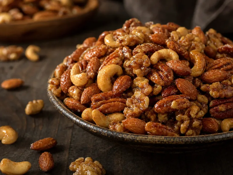 Eine Schale glänzend ahornglasierter gemischter Nüsse — Walnüsse, Pekannüsse, Mandeln und Cashews — karamellisiert und mit feinen Zuckerkristallen überzogen auf einem dunklen Holztisch