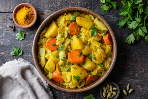 Schale mit äthiopischem Atakilt Wat: gelb leuchtende Kartoffelstücke, Karottenscheiben und Weißkohlstreifen mit Kurkuma, garniert mit frischer Petersilie, auf dunklem Holztisch