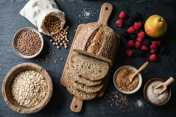 Ballaststoffreiche pflanzliche Lebensmittel in moodiger Stillleben-Anordnung — Haferflocken, Linsen, Kichererbsen, Vollkornbrot, Beeren, Birne, Leinsamen und Flohsamenschalen auf dunklem Untergrund