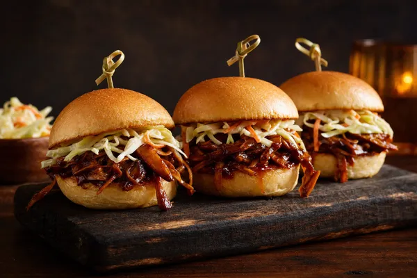 Drei kleine Slider-Burger mit zerzupften, glänzend BBQ-glasierten Pilzen und hellem Krautsalat auf weichen Brötchen, aufgespießt und gestapelt auf einem Holzbrett