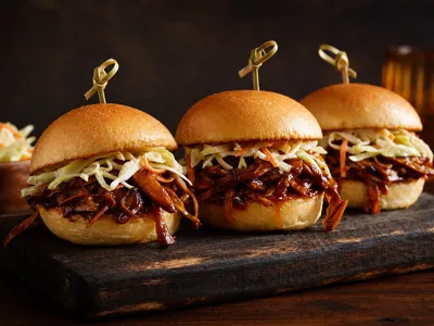 Drei kleine Slider-Burger mit zerzupften, glänzend BBQ-glasierten Pilzen und hellem Krautsalat auf weichen Brötchen, aufgespießt und gestapelt auf einem Holzbrett