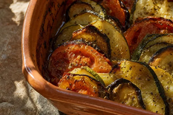 Nahaufnahme einer Tonform mit griechischem Briam: dachziegelartig geschichtete Scheiben von Zucchini, Aubergine, Kartoffel und Tomate mit dunkel gerösteten Rändern, glänzend von Olivenöl und Oregano