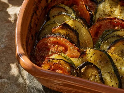 Nahaufnahme einer Tonform mit griechischem Briam: dachziegelartig geschichtete Scheiben von Zucchini, Aubergine, Kartoffel und Tomate mit dunkel gerösteten Rändern, glänzend von Olivenöl und Oregano