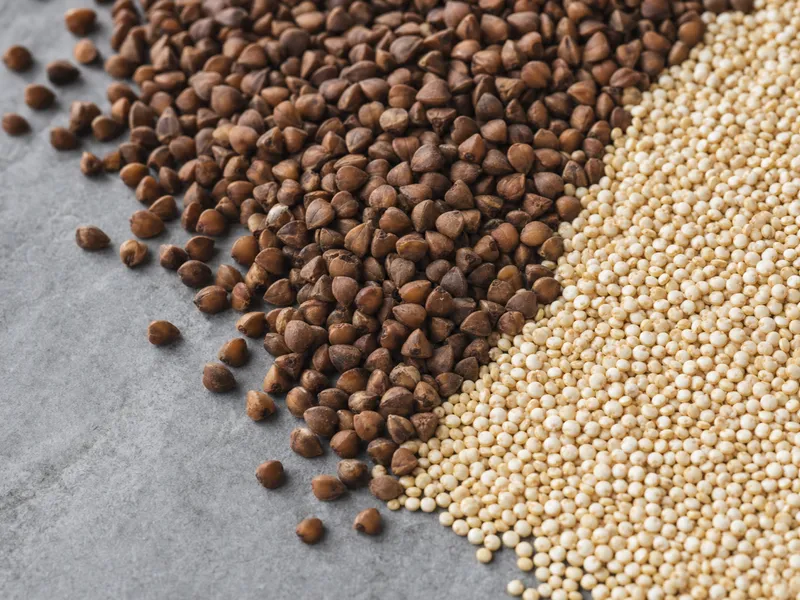 Nahaufnahme zweier Getreidehaufen auf grauem Stein: dunkle Buchweizenkörner links, helle goldene Quinoa-Körner rechts, an einer diagonalen Grenze aufeinandertreffend