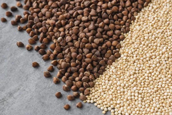 Nahaufnahme zweier Getreidehaufen auf grauem Stein: dunkle Buchweizenkörner links, helle goldene Quinoa-Körner rechts, an einer diagonalen Grenze aufeinandertreffend