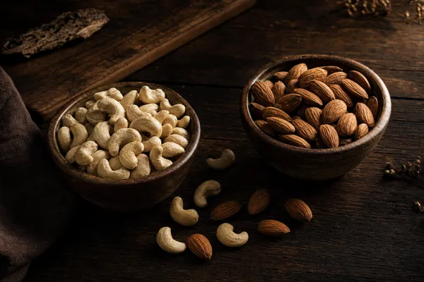 Zwei Keramikschalen auf dunklem Holztisch: links helle Cashewkerne, rechts braune Mandeln, dazwischen einzelne verstreute Nüsse
