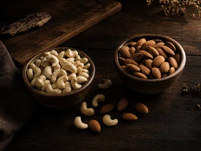 Zwei Keramikschalen auf dunklem Holztisch: links helle Cashewkerne, rechts braune Mandeln, dazwischen einzelne verstreute Nüsse