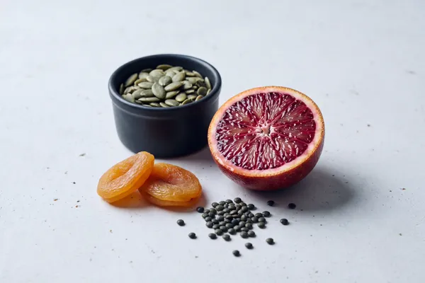 Stillleben mit Kürbiskernen in einer dunklen Schale, getrockneten Aprikosen, grünen Linsen und einer halbierten Blutorange auf hellem Stein — pflanzliche Eisenquellen mit Vitamin-C-Begleiter