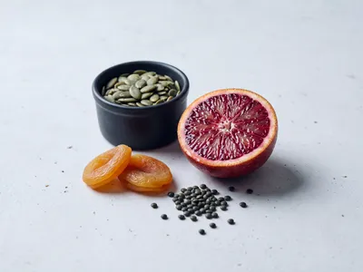 Stillleben mit Kürbiskernen in einer dunklen Schale, getrockneten Aprikosen, grünen Linsen und einer halbierten Blutorange auf hellem Stein — pflanzliche Eisenquellen mit Vitamin-C-Begleiter