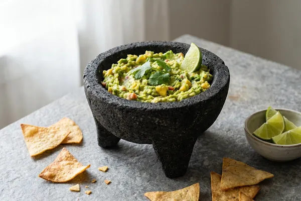 Steinmörser (Molcajete) mit frisch gestampfter Guacamole, garniert mit Koriander und Limettenspalten, daneben goldbraun gebackene Totopos-Chips auf dunklem Steintisch