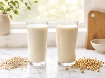 Zwei Gläser cremiger Pflanzenmilch auf heller Marmorplatte: daneben links ein Häufchen Haferflocken, rechts getrocknete Sojabohnen