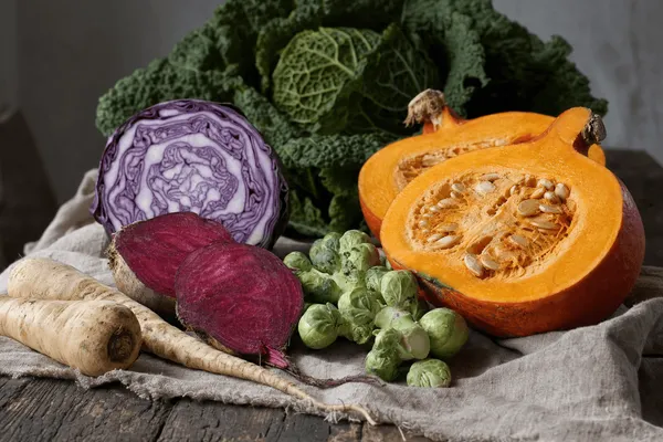 Herbstgemüse auf grobem Leinen auf einem alten Holztisch: ein halbierter Hokkaido-Kürbis mit Kernen, ein angeschnittener Rotkohl mit violett-weißer Maserung, halbierte Rote Bete, Rosenkohl und Pastinaken, dahinter ein Wirsing