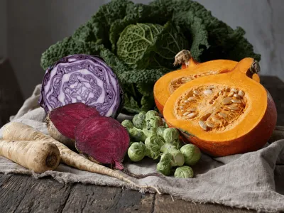 Herbstgemüse auf grobem Leinen auf einem alten Holztisch: ein halbierter Hokkaido-Kürbis mit Kernen, ein angeschnittener Rotkohl mit violett-weißer Maserung, halbierte Rote Bete, Rosenkohl und Pastinaken, dahinter ein Wirsing