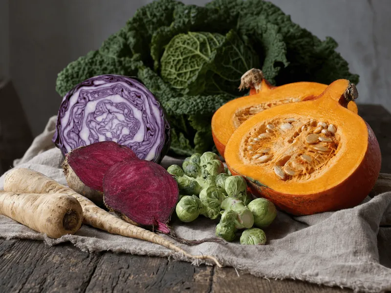 Herbstgemüse auf grobem Leinen auf einem alten Holztisch: ein halbierter Hokkaido-Kürbis mit Kernen, ein angeschnittener Rotkohl mit violett-weißer Maserung, halbierte Rote Bete, Rosenkohl und Pastinaken, dahinter ein Wirsing