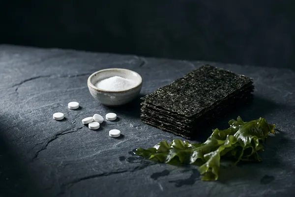 Pflanzliche Jodquellen für vegane Ernährung — Nori-Algenblätter, jodiertes Speisesalz im Schälchen und Jod-Tabletten in moodiger Stillleben-Anordnung