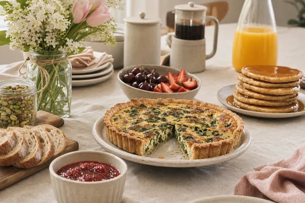 Frühlings-Brunchtisch mit pflanzlicher Spinat-Tofu-Quiche, Sauerteig-Brot, Erdbeer-Chia-Marmelade, veganen Pancakes mit Ahornsirup, frischen Erdbeeren und Kirschen, einer Karaffe Orangensaft und einem Strauß Tulpen — Muttertags-Brunch ohne Bekenntnis-Geste
