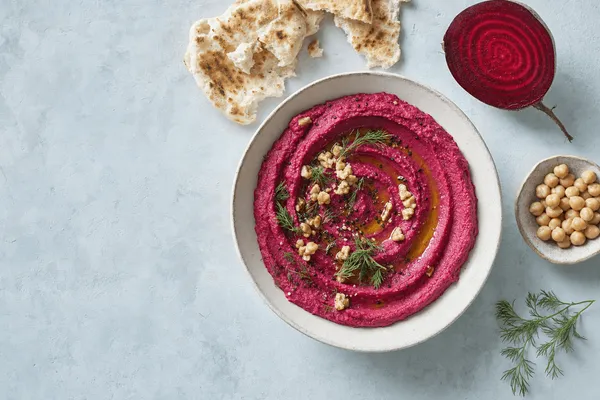Draufsicht auf eine flache helle Keramikschale mit leuchtend pinkem Rote-Bete-Hummus, in Spiralen gestrichen, mit Olivenöl, gehackten Walnüssen und Dill, daneben Fladenbrot und eine halbierte Rote Bete auf blaugrauem Putz