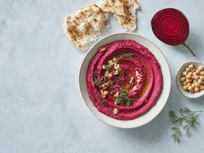 Draufsicht auf eine flache helle Keramikschale mit leuchtend pinkem Rote-Bete-Hummus, in Spiralen gestrichen, mit Olivenöl, gehackten Walnüssen und Dill, daneben Fladenbrot und eine halbierte Rote Bete auf blaugrauem Putz