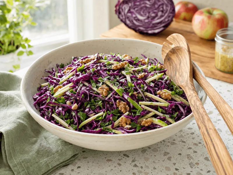 Cremefarbene Keramikschüssel mit fein gehobeltem violettem Rotkohl, grünen Apfelstiften, gerösteten Walnusshälften und Petersilie, daneben Salatbesteck aus Olivenholz auf einer hellen Terrazzo-Arbeitsplatte im Morgenlicht
