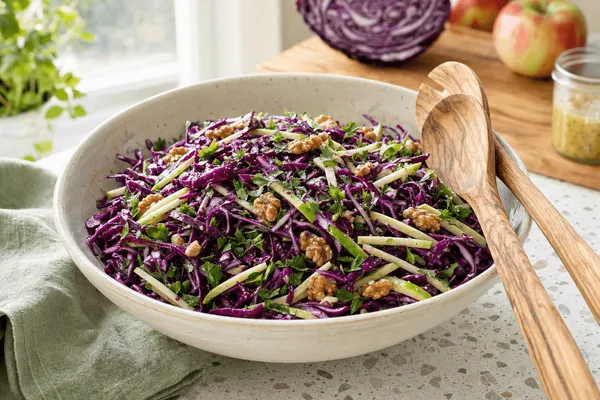 Cremefarbene Keramikschüssel mit fein gehobeltem violettem Rotkohl, grünen Apfelstiften, gerösteten Walnusshälften und Petersilie, daneben Salatbesteck aus Olivenholz auf einer hellen Terrazzo-Arbeitsplatte im Morgenlicht