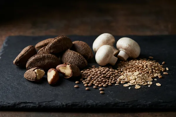Pflanzliche Selenquellen — aufgebrochene Paranüsse, Champignons, Linsen und Vollkorn auf dunklem Schiefer in moodiger Stillleben-Anordnung