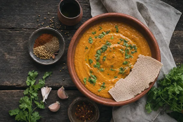 Rustikale Tonschale mit cremigem äthiopischem Shiro aus Kichererbsenmehl, ockerfarben und glänzend, garniert mit frischem Koriander, daneben ein Stück Injera auf dunklem Holztisch