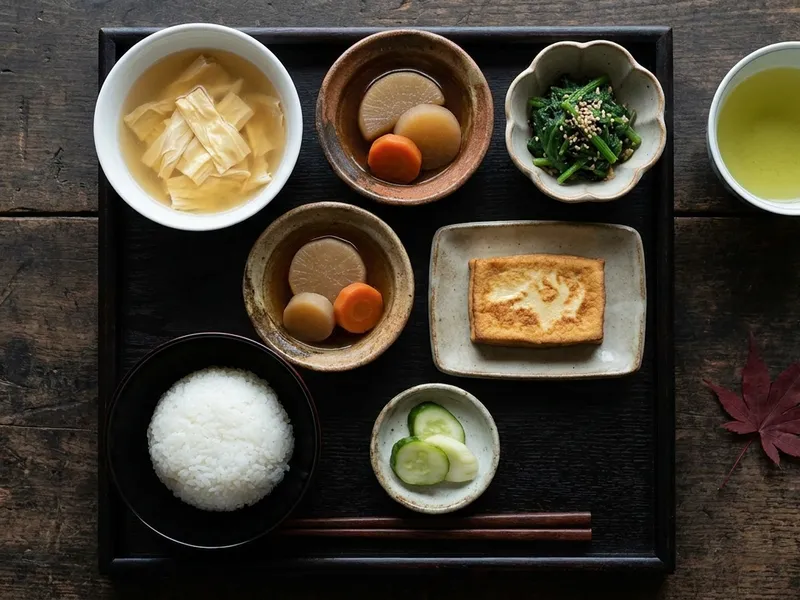 Traditionelles Shōjin-Ryōri-Tablett mit Miso-Suppe, Reis, gedünstetem Daikon, Sesam-Spinat, Koyadofu und grünem Tee auf dunklem Holz