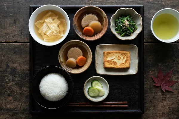 Traditionelles Shōjin-Ryōri-Tablett mit Miso-Suppe, Reis, gedünstetem Daikon, Sesam-Spinat, Koyadofu und grünem Tee auf dunklem Holz