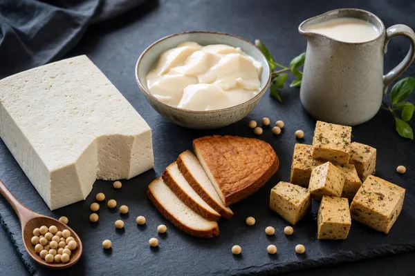 Moodiges Stillleben auf dunklem Schiefer: nebeneinander ein Block fester Naturtofu, eine Schale cremiger Seidentofu, in Scheiben geschnittener brauner Räuchertofu und marinierte Tofuwürfel, dazu Sojabohnen, eine Sojamilchkanne und ein Holzlöffel.