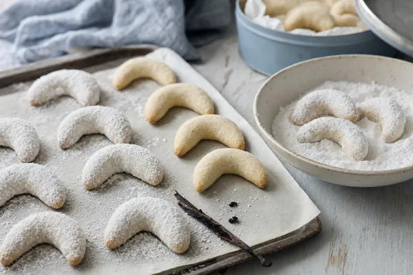Frisch gebackene vegane Vanillekipferl auf einem Backblech mit Backpapier, ein Teil dick mit Vanille-Puderzucker überzogen, daneben eine Schale Vanillezucker mit drei Kipferln und eine ausgekratzte Vanilleschote auf einem graublauen Holztisch