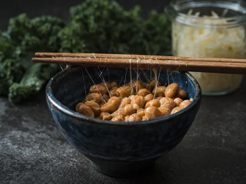Natto in einer indigoblau glasierten Keramikschale, zwischen den Sojabohnen und den aufgelegten Holzstäbchen ziehen sich feine Fäden, im unscharfen Hintergrund Grünkohl und ein Glas Sauerkraut