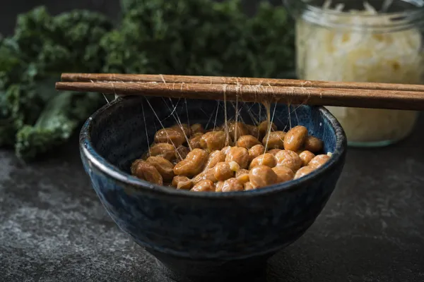Natto in einer indigoblau glasierten Keramikschale, zwischen den Sojabohnen und den aufgelegten Holzstäbchen ziehen sich feine Fäden, im unscharfen Hintergrund Grünkohl und ein Glas Sauerkraut