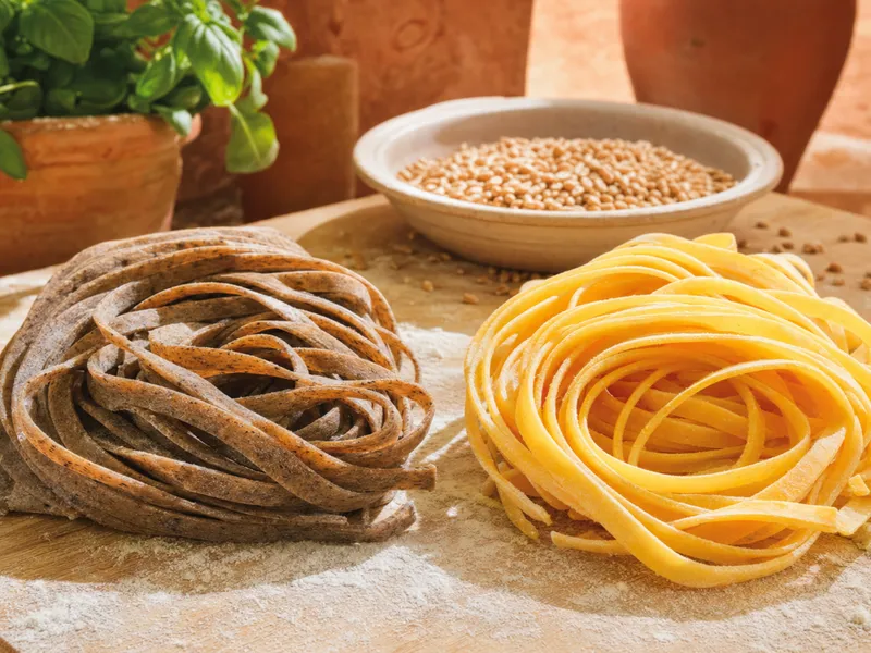 Zwei Nester getrockneter Tagliatelle auf bemehltem Holzbrett: dunkle Vollkornnudeln links, helle Semolina-Nudeln rechts, dahinter eine Schale Weizenkörner