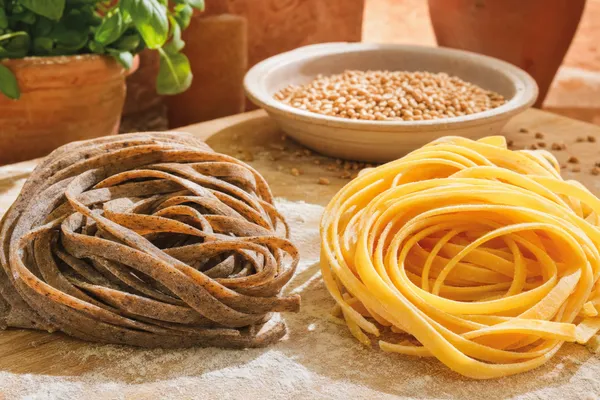 Zwei Nester getrockneter Tagliatelle auf bemehltem Holzbrett: dunkle Vollkornnudeln links, helle Semolina-Nudeln rechts, dahinter eine Schale Weizenkörner