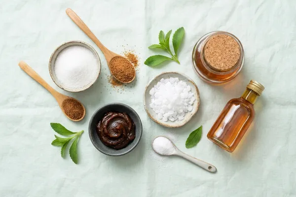 Verschiedene Zuckerersatzstoffe nebeneinander: Stevia-Blätter, Erythrit-Kristalle, Xylit-Pulver, Dattelpaste, Reissirup und Ahornsirup auf hellem Leinen-Hintergrund